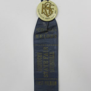 May include: Blue ribbon with gold lettering that reads "Kentucky State Fair Louisville First Premium" and "Vegetables and Melons". A white button with a gold "K.S.F." logo and the text "Louisville Sept. 14th 1908" is attached to the top of the ribbon.