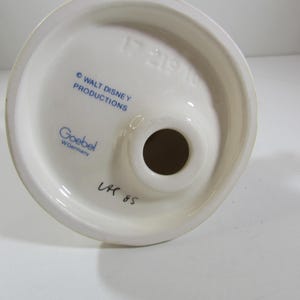 May include: A white ceramic figurine base with a circular top and a central hole. The base features blue text that reads "&copy; WALT DISNEY PRODUCTIONS" and "Goebel W. Germany". Handwritten text "LK 85" is also visible.