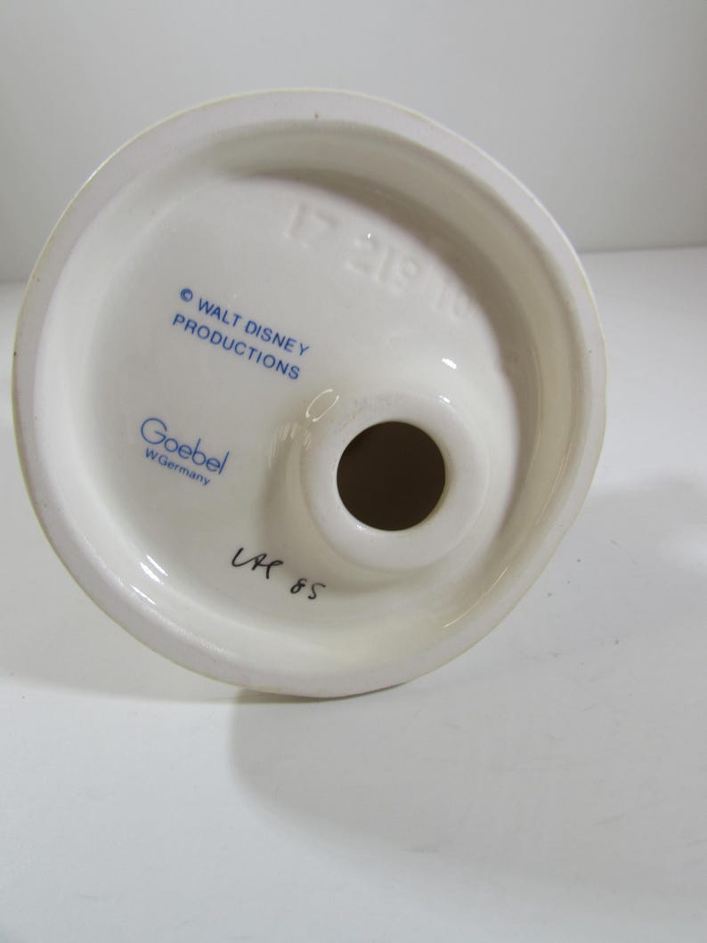 May include: A white ceramic figurine base with a circular top and a central hole. The base features blue text that reads "&copy; WALT DISNEY PRODUCTIONS" and "Goebel W. Germany". Handwritten text "LK 85" is also visible.