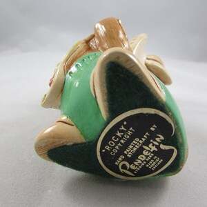 May include: A hand-painted Pendelfin stonecraft figurine of "Rocky" in a seated position. The figurine features a green vest, beige accents, and a dark green base. The base has a black and white label with text.