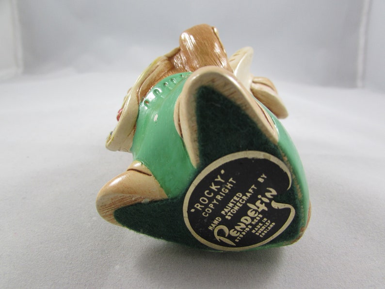 May include: A hand-painted Pendelfin stonecraft figurine of "Rocky" in a seated position. The figurine features a green vest, beige accents, and a dark green base. The base has a black and white label with text.