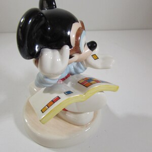 May include: A ceramic figurine of a cartoon mouse with black ears, a blue shirt, and white gloves, reading a book with colorful squares. The figurine is on a round, off-white base.