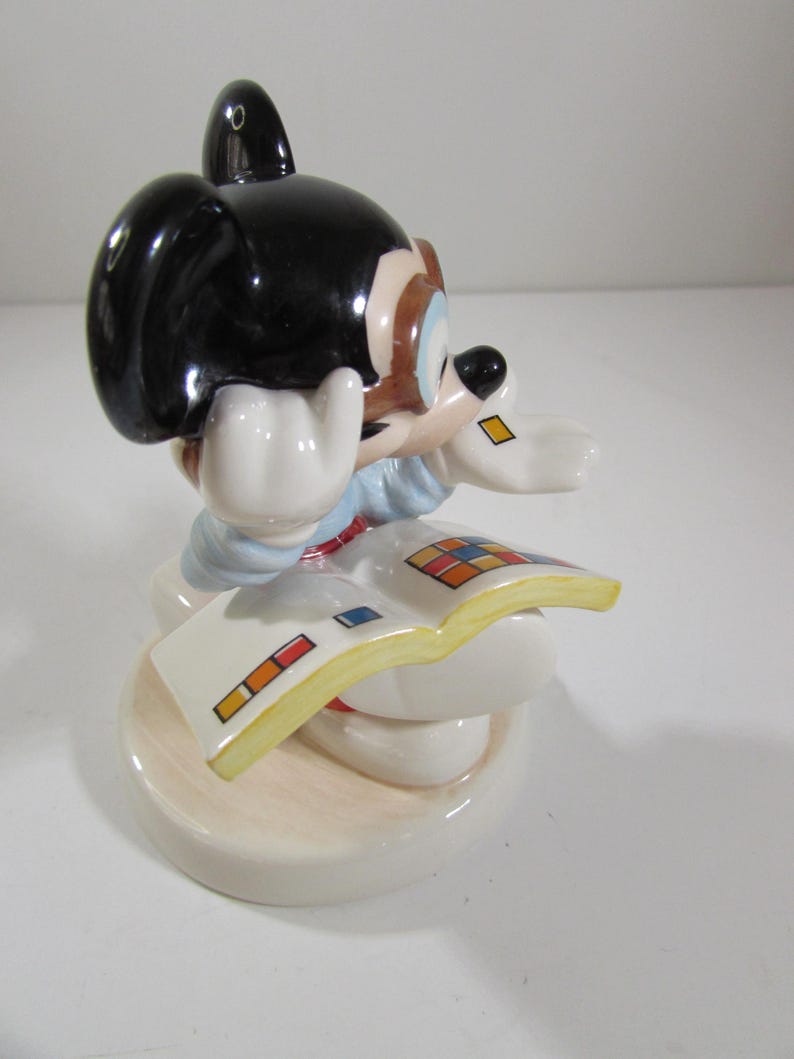May include: A ceramic figurine of a cartoon mouse with black ears, a blue shirt, and white gloves, reading a book with colorful squares. The figurine is on a round, off-white base.