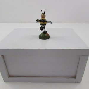 May include: A white wooden box with a small wooden bee figurine on top. The bee is painted yellow and black with a black and white face. The bee is standing on a green base.