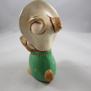May include: A ceramic figurine of a bunny wearing a hat and green shorts. The bunny is in a kneeling position, with its hands clasped together. The figurine is painted in shades of beige, brown, and green.