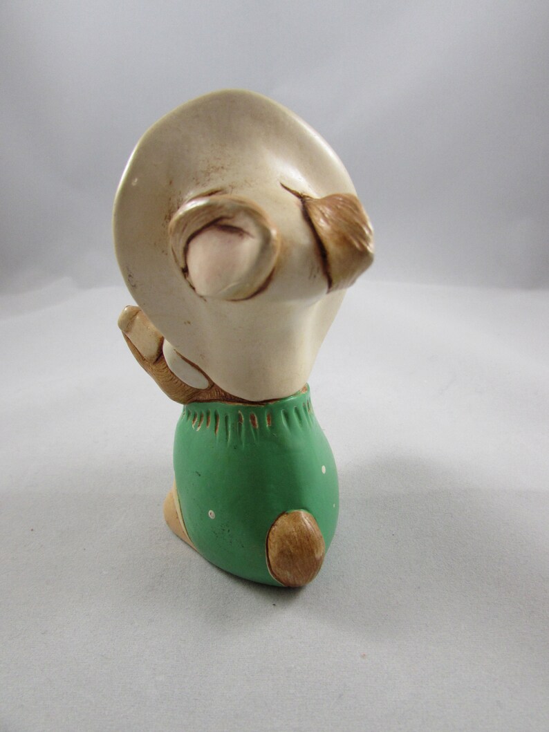 May include: A ceramic figurine of a bunny wearing a hat and green shorts. The bunny is in a kneeling position, with its hands clasped together. The figurine is painted in shades of beige, brown, and green.