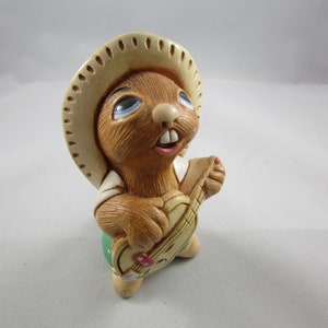 May include: A ceramic figurine of a brown rabbit wearing a sombrero and playing a small guitar. The rabbit has blue eyes and is dressed in a white shirt and green pants. The guitar has a red and white design.
