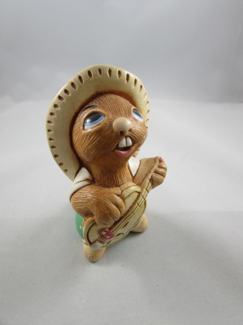 May include: A ceramic figurine of a brown rabbit wearing a sombrero and playing a small guitar. The rabbit has blue eyes and is dressed in a white shirt and green pants. The guitar has a red and white design.