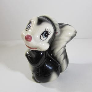 May include: A black and white ceramic figurine of a skunk with a pink nose and large blue eyes. The skunk is standing on all fours and has a white stripe down its back.