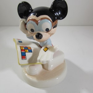 May include: A ceramic figurine of Mickey Mouse wearing glasses and holding a paper with colorful squares. The figurine has black ears, a black nose, and is wearing a light blue shirt. The base is off-white with a light brown accent.