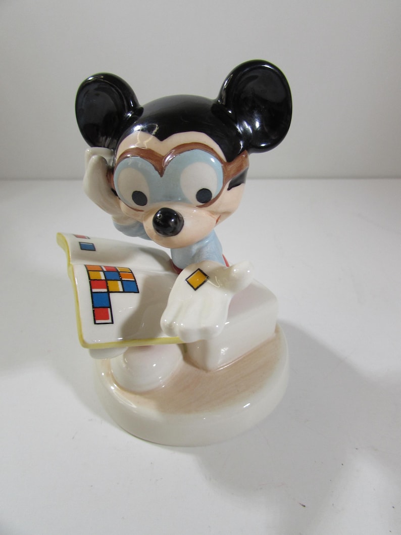 May include: A ceramic figurine of Mickey Mouse wearing glasses and holding a paper with colorful squares. The figurine has black ears, a black nose, and is wearing a light blue shirt. The base is off-white with a light brown accent.