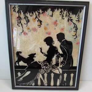 May include: A framed silhouette art piece featuring a black cutout of a couple and a small dog sitting on a balcony railing. The background is a textured cream material decorated with small, colorful fabric flowers and black hanging vines.