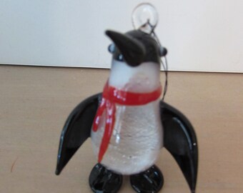 May include: A glass penguin ornament with black wings, a white body, and a red scarf. The ornament has a clear glass loop for hanging.
