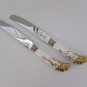 May include: Two silver butter knives with white porcelain handles decorated with gold grapevine designs.