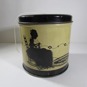 May include: A vintage metal tin with a black lid and a beige body. The tin features a black silhouette of a woman sitting in a rocking chair knitting. The tin is designed to hold yarn.