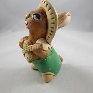 May include: A ceramic figurine of a brown rabbit wearing a beige sombrero and green shorts. The rabbit is holding a basket. The figurine has blue eyes and is posed as if it is looking up.