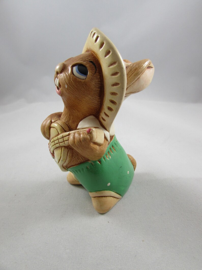 May include: A ceramic figurine of a brown rabbit wearing a beige sombrero and green shorts. The rabbit is holding a basket. The figurine has blue eyes and is posed as if it is looking up.