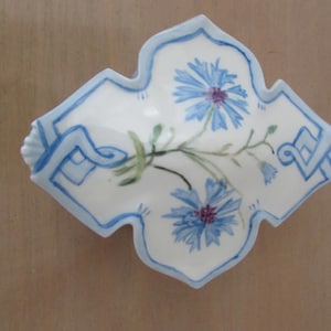 May include: A white ceramic dish with a blue and white floral design. The dish is shaped like a diamond and has a scalloped edge. The flowers are blue and have a pink center.