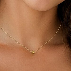 May include: A gold-toned necklace featuring a delicate chain and a single, small, square-shaped bead charm centered on the collarbone. The charm is engraved with the letter M on its front face, providing a subtle and refined detail to the piece.