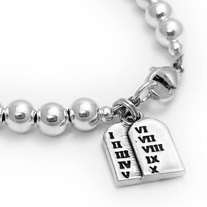 May include: A silver bracelet with a silver clasp and two silver charms shaped like tablets with Roman numerals inscribed on them. The bracelet is made of small silver beads.