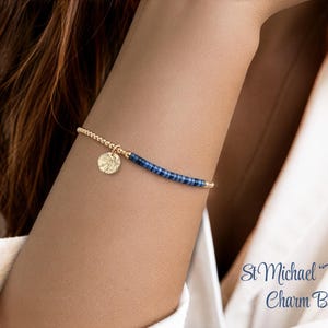 May include: A beaded bracelet featuring a gold-toned circular charm with a textured surface, small gold-toned beads, and a section of small dark blue cylindrical beads. The text St Michael Protect Us Charm Bracelets is visible in the corner.