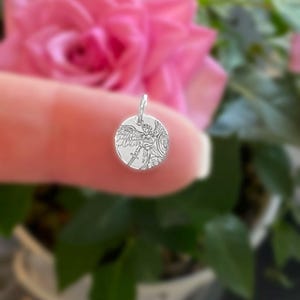 May include: A small, round silver pendant featuring an engraved image of an angel with wings, a sword, and a shield. The pendant is held up against a blurred background of a pink rose and green foliage.
