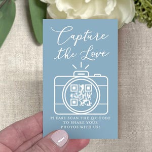 May include: A light blue card with the text "Capture the Love" in cursive script. Below the text is a white camera graphic with a QR code. The card also includes the text "PLEASE SCAN THE QR CODE TO SHARE YOUR PHOTOS WITH US!"