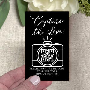 May include: A black rectangular card with white text and a camera graphic. The text reads "Capture the Love" and "PLEASE SCAN THE QR CODE TO SHARE YOUR PHOTOS WITH US!". A QR code is in the center of the camera graphic.