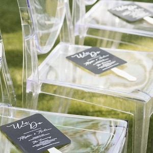 May include: Clear acrylic chairs arranged on green grass, each with a black fan featuring white text that reads "We Do" and names. The chairs have a modern, minimalist design, suitable for outdoor events.