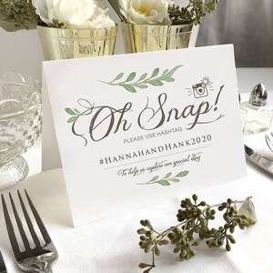 May include: A white card with green foliage and a camera graphic. The text on the card reads "Oh Snap! Please use hashtag #HANNAHANDHANK2020 To help us capture our special day."