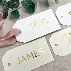 May include: Three white gift tags with gold lettering. The tags have a hole punched in the top left corner. The tags are labeled "Kelly", "JAMES", and "G".