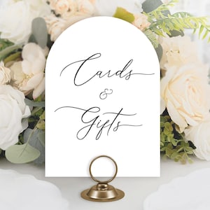 May include: White arch-shaped card with black calligraphy that reads "Cards & Gifts" on a white table with a gold metal stand. The card is surrounded by white and cream flowers.