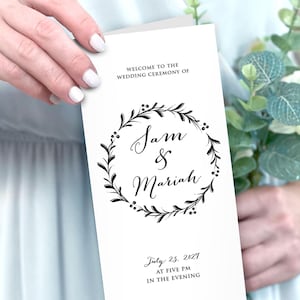 May include: A white wedding ceremony program featuring a black ink botanical wreath graphic surrounding the names Sam & Mariah. The text includes the date July 25, 2027, at five pm in the evening, and the location Riverside Farm, Pittsfield, Vermont.