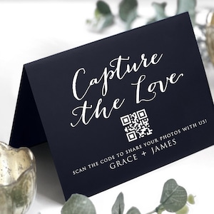 May include: A navy blue folded card with the text "Capture the Love" in white script. A QR code and the text "SCAN THE CODE TO SHARE YOUR PHOTOS WITH US! GRACE + JAMES" are also visible. The card is on a white surface.