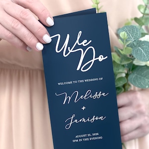 May include: A navy blue wedding program with white calligraphy that reads "We Do" and "Welcome to the Wedding of Melissa + Jamison". The program also includes the date and time of the wedding, August 22, 2026, 5 PM in the evening, and the location, Maple Farms, Austin, Texas.
