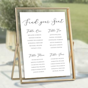 May include: A gold-framed seating chart with the words "Find your Seat" in elegant script. The chart lists names under "Table One" through "Table Four". The background is blurred, suggesting an outdoor setting.