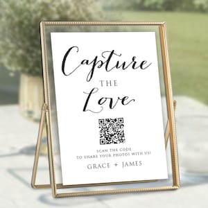 May include: A gold-framed sign with the words "Capture the Love" in elegant script. Below the text is a QR code and the message "Scan the code to share your photos with us!" followed by the names "Grace + James". The sign is displayed on a table.