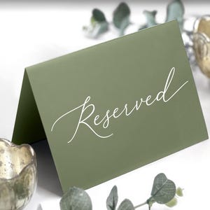 May include: A folded olive green card with the word "Reserved" in elegant white script. The card is positioned on a white surface, with decorative elements including a gold-toned candle holder and greenery.