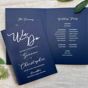 May include: Navy blue wedding program with white text. The front cover says "We Do" and includes the names Gianna and Christopher, the date August 18, 2023, and the location Westhampton Beach, New York. The back cover lists the wedding party, including the officiant, bridesmaids, groomsmen, flower girl, ring bearer, and ushers.