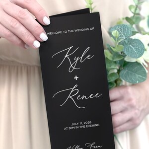 May include: Black wedding program with white text. The program says "Welcome to the wedding of Kyle + Renee" and includes the date "July 11, 2026" and the location "Valley Farm, Ashland, Virginia".