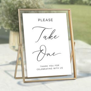 May include: A gold-framed sign with the words "PLEASE Take One" in elegant script. The sign also includes the text "THANK YOU FOR CELEBRATING WITH US". The sign is displayed on a white surface, with a blurred green background.