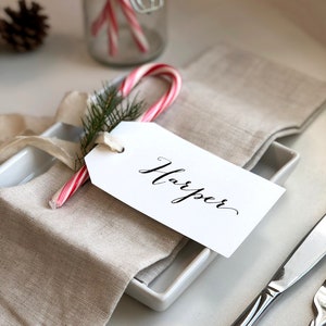 May include: A white gift tag with the name "Harper" written in black calligraphy, tied to a red and white candy cane with a sprig of green pine. The tag is placed on a folded linen napkin, next to a white square plate and silver cutlery.