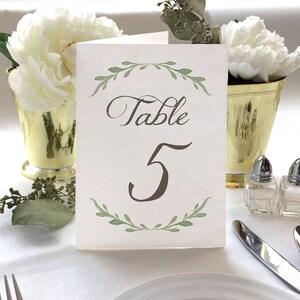 May include: A white card with green foliage border and the text "Table 5" in brown calligraphy.