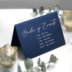 May include: A navy blue card with white text that reads "Order of Events" and lists the order of events for a wedding reception with times. The events include: 4:00 We Do, 5:00 We Drink, 7:00 We Eat, 8:00 We Dance, 10:00 We Leave.