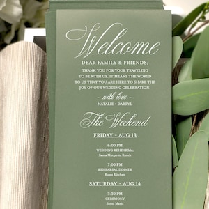May include: A stack of green wedding itinerary cards with white script. The cards list the wedding weekend schedule, including the ceremony and reception times. The top card reads "Welcome" in elegant script.