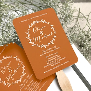 May include: Orange wedding ceremony program fans with white text. The fans feature a floral wreath design and the names Olivia & Michael. The text includes the date, time, and order of events for the ceremony and reception.