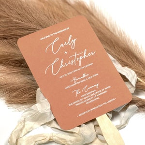 May include: A peach-colored wedding fan with white text that reads "Welcome to the wedding of Carly + Christopher July 22, 2022 | 5PM in the evening Durabler Westhampton Beach, New York The Ceremony Processional Exchange of vows | Exchange of rings Pronouncement of marriage Recessional Reception immediately following."