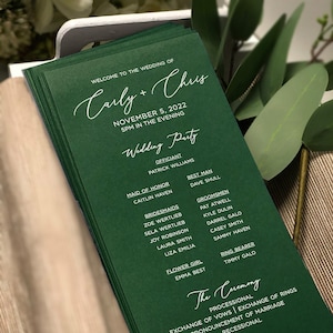 May include: Green wedding program cards with white text. The cards list the wedding party members and the order of the ceremony. The text reads "Welcome to the wedding of Carly + Chris, November 5, 2022, 5PM in the evening, Wedding Party, Officiant, Patrick Williams, Maid of Honor, Caitlin Haven, Bridesmaids, Zoe Wertlieb, Sela Wertlieb, Joy Robinson, Laura Smith, Liza Emilia, Flower Girl, Emma Best, Best Man, Dave Shull, Groomsmen, Pat Atwell, Kyle Dulin, Darrel Galo, Casey Smith, Sammy Haven, Ring Bearer, Timmy Galo, The Ceremony, Processional, Exchange of Vows | Exchange of Rings, Pronouncement of Marriage, Recessional, Thank you for sharing this special day with us."