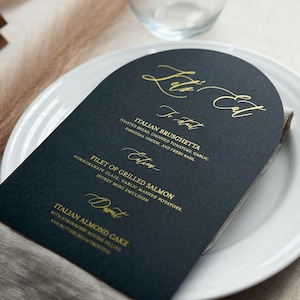 May include: An arched dark navy blue menu card with gold foil lettering. The text reads Let's Eat, To Start, Italian Bruschetta, Entrees, Filet of Grilled Salmon, Dessert, and Italian Almond Cake, along with descriptions of the food items.