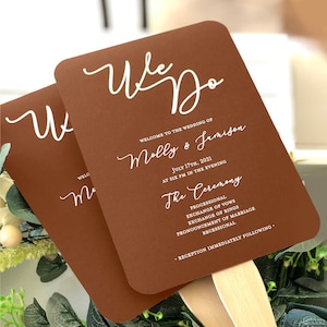 May include: Two brown wedding fans with wooden handles. The fans have rounded corners and white script that reads "We Do" and wedding details for Molly & Jamison, including the date and ceremony schedule.
